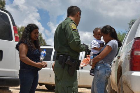 One woman holding an infant and another woman with dark hair stand next to a border patrol agent wearing a green uniform