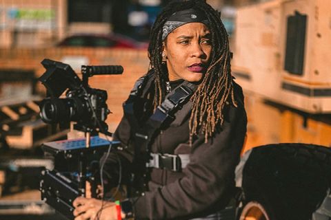 Firelight. Black person with long brown locs wearing black clothing with a black headband, holding  a large camera.