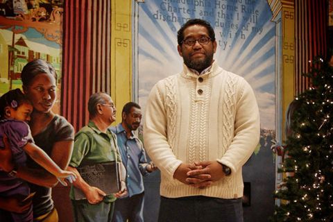 James Hough. Black man with short dark hair and beard wearing cream colored sweater, jeans, standing in front of a mural.