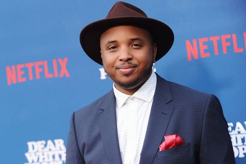Justin Simien. Black man wearing brown fedora, blue suit jacket and white shirt, standing in front of Netflix signage.