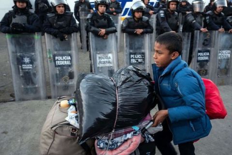Young boy pushes his belongings in a grocery cart as he walks past a long line of Mexican officers 
