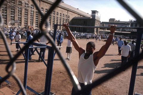 Black man in beige prison uniform does pull-ups outside in a prison yard. 