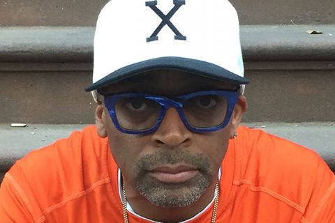 Spike Lee. Black man wearing white "X" cap with blue glasses and orange t-shirt.