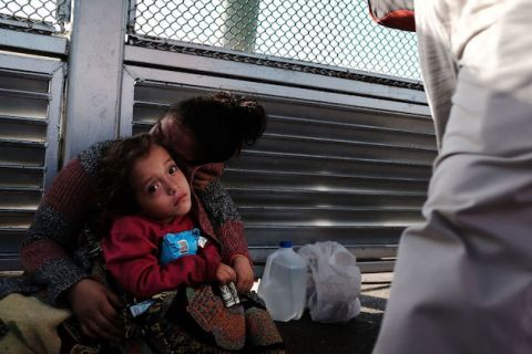 A small child sits on mother's lap as they wait in front of a metal fence. 