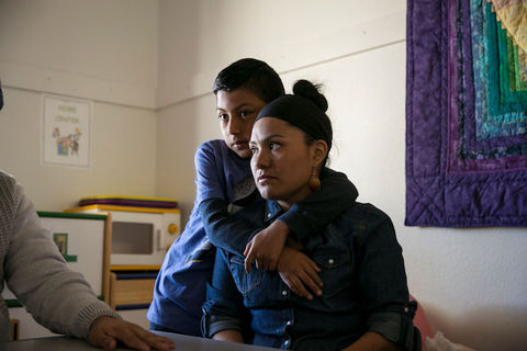 Latinx woman wearing denim shirt sits as her young son stands behind her with his arms wrapped around her shoulders. 