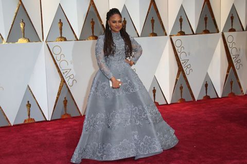 Ava DuVernay. Black woman with long brown locs wearing silver gown on red carpet standing in front of Oscar statues.