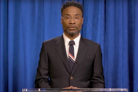 Billy Porter. Black man with short dark hair wearing dark suit jacket, blue and red tie with white shirt and glasses, standing in front of blue curtain.