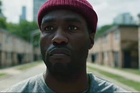 Yahya Abdul-Mateen II. Black man wearing dark red wool skull cap and green t-shirt standing in front of a row of buildings.