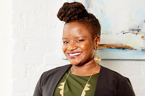 Chinyere Ezie. Smiling Black woman with locs in a high bun and shaved sides, wearing a green shirt, dark suit jacket and gold earrings and necklace.