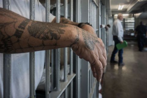 Man with tattoos on his hands and forearms hangs his hands through jail cell bars. 