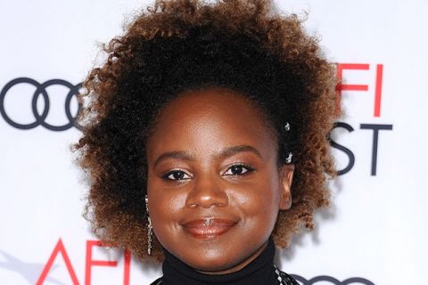 Dee Rees. Black woman with brown Afro, smiling, wearing a dark top in front of a step and repeat sign.