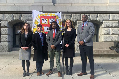 Harvard students. Five students stand side-by-side. Two Black men wearing gray suits, one White woman wearing black jacket and gray dress, Asian American woman wearing all black dress and black jacket, Latinx person wearing black coat, colorful tie, white shirt.