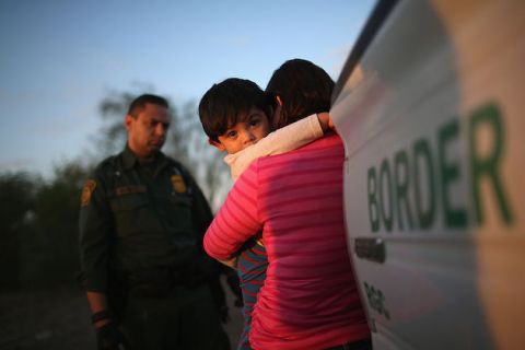 A small, dark-haired boy hugs his mother tightly as she holds him and wraps him in her arms. She stands near a truck marked BORDER.