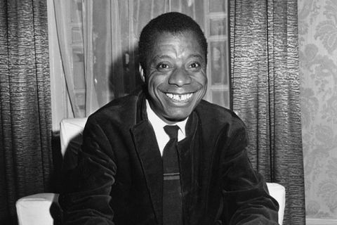 James Baldwin. Black man with short dark hair smiling broadly, wearing a white shirt and dark suit jacket, with dark tie.