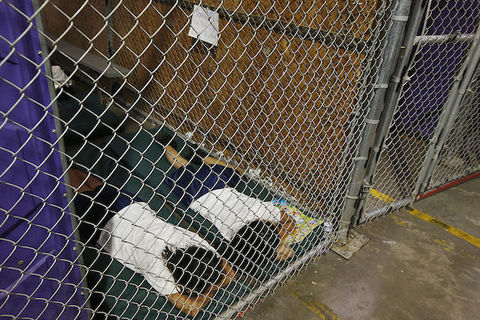 Two people wearing white t-shirts lie face down on a black mat inside of a wire cage inside an Arizona immigration detention center.
