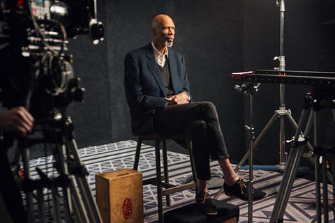 Kareem Abdul Jabbar. Slim older Black man with bald head and gray beard, wearing blue blazer, gray V-neck sweater and pants and a white button down top. He's sitting in chair with legs crossed surrounded by camera equipment.