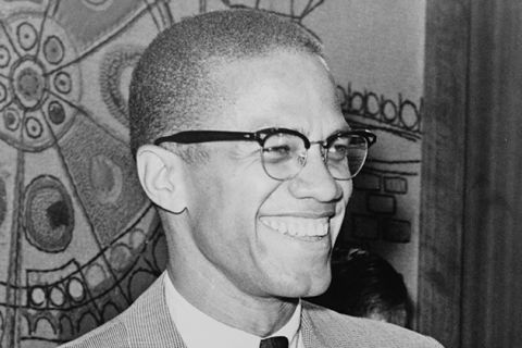 Malcolm X. Black man with short brown hair wearing glasses, white shirt, black tie, tan colored suit jacket, smiling.