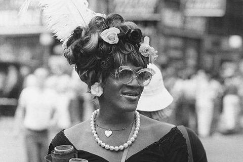 Marsha P. Johnson. Black woman with flowers and feather in hair, wearing glasses, flower earrings, pearl necklace and black sweetheart shaped top.