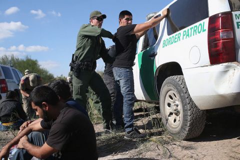 Man stands with his back to border patrol agent as border agent frisks him up against a white border patrol van.
