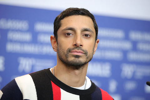Riz Ahmed. South Asian man with short dark hair and short, groomed facial hair wearing red, white, blue colored sweater.