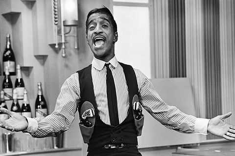 Sammy Davis Jr. Black man with short dark hair singing in a gun-slinger's outfit seated on a bar with his arms extended.