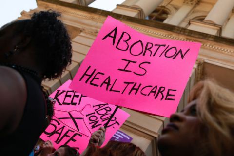 A pink protest sign that reads "Abortion is healthcare" is held at a rally in front of a government building