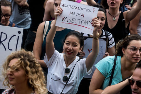 An Asian woman in a white t-shirt is at a protest and holds a sign that reads "Against abortion? Have a vasectomy."