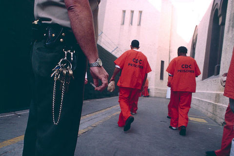 a group of men wearing orange jumpsuits walk away from the camera as a security officer can be seen in the foreground.