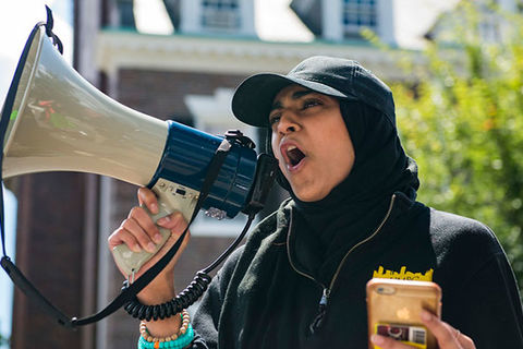 Anti-Hate Report. Muslim woman wearing a black cap over a black headscarf and black top holding a bullhorn.