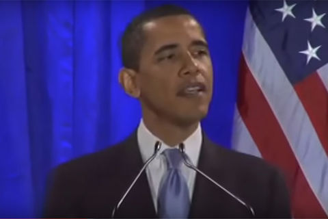 Barack Obama. Black man with short dark hair wearing a dark suit jacket, blue tie and white shirt standing in front of a blue curtain and the American flag.