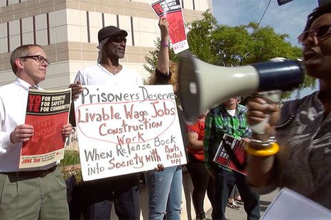 Scene from the documentary 'Bedlam' :  Protest scene where two men, one White, one Black, hold up signs and Black woman walks by speaking on a microphone.