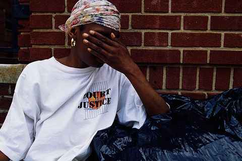 COVID-19-Domestic Violence. Young Black woman looking distressed in front of a brick building covers her face, wearing a colorful headscarf and white T-shirt, standing behind black garbage bags.