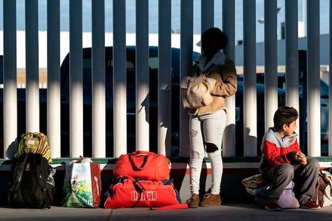A parent and child hold all their belongings and lean against a large, white fence. 