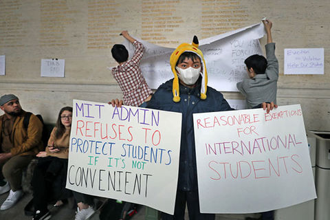 Coronavirus protest. Asian-American student wearing yellow hat, white face mask, blue coat holding up to two protest signs.