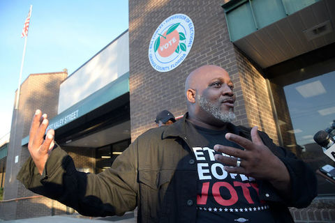 Black man wears a t-shirt that reads, "let my people vote" as he stands in front of a brick building.