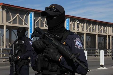 Border patrol agents wear black masks over their entire faces and hold guns while standing in the street. 