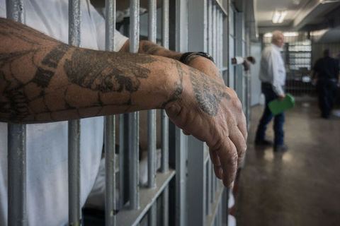 a man with tattoos hangs his arms through the bars of a prison cell. 