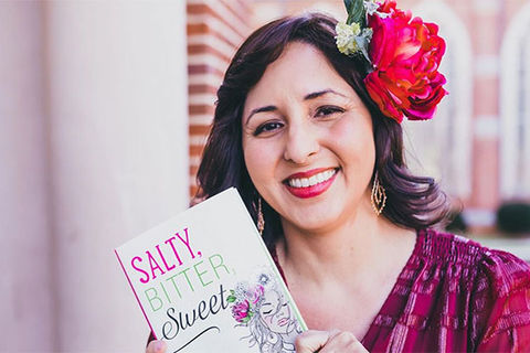 Mayra Cueva. Latinx person with a red flower in their hair, wearing a red top and holding up a children's book.