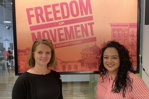 Two women stand in front of an orange sign that reads "Freedom of Movement"