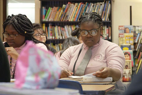 PUSHOUT. Two teenage Black girls in a classroom with book shelves behind them as they sit at a desk wearing pink button up shirts, gray ties, long braids and glasses. 