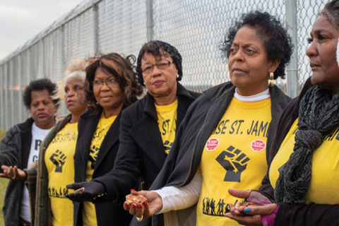 RISE activists. Six older Black women standing side-by-side against a fence holding a hand out, all wearing yellow t-shirts and black jackets.