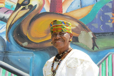 Sadie Roberts Joseph. Older Black woman wearing colorful hat with white top and brown beaded necklace, standing in front of colorful wall mural.