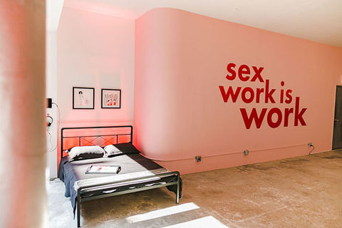 Sex Workers Popup. Pink wall reads "sex work is work" next to a bed.