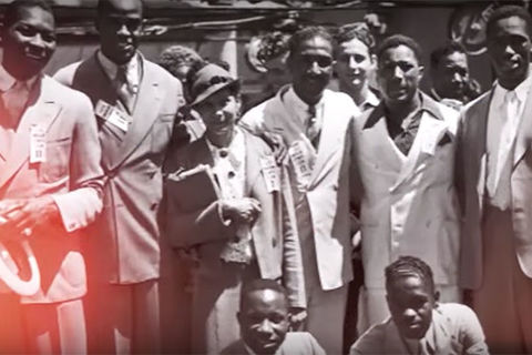 1936 Olympics. Archival image of about nine Black men and women huddled together for a photo with everyone wearing a suit.