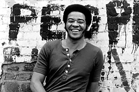 Bill Withers. Black and white photo with Black man in a short Afro and dark short sleeve t-shirt standing in front of brick wall with "People" spray painted on it.