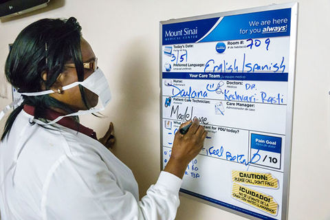 Black nurse. Black woman with dark hair wearing a white mask and lab coat writing at a schedule board.