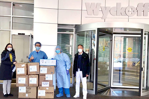 Four people standing in front of Wycoff Hospital and around many brown boxes.