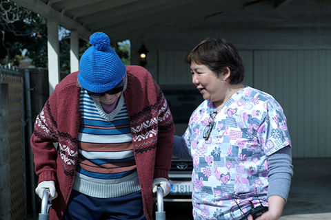 “COVER/AGE”. Older Asian American woman in floral print scrubs assisting an elderly person using a walker.