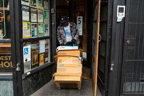 COVID-19 NYC 2020. Black man wearing dark Yankees cap , T-shirt and plaid jacket pulling boxes into the doorway of a shop. 