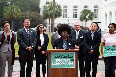 Campaign for Southern Equality. Black woman with Afro wearing a dark suit and classes stands outside in front of wooden podium, in front a row of people, also in suits.